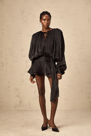 Model wearing the Evelyn Black Draped Mini Dress, showcasing the fully pleated design, twist waist detail, and elegant long buttoned-cuff sleeves.Black Little Dress Ruffled Mini Longsleeve