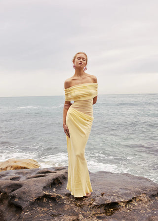 Woman in a yellow strapless gown standing on rocks by the ocean.