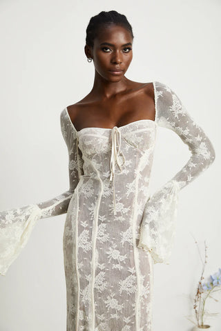Elegant woman wearing a white bohemian lace maxi dress with long bell sleeves, intricate floral lace pattern, and a sweetheart neckline, standing gracefully in a minimalistic setting with a small vase of flowers. - Cielie Vienna