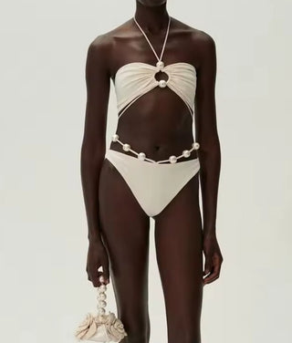 Woman wearing a pearl-accented ivory bikini set with criss-cross halter straps and high-cut bottoms in a tropical garden setting