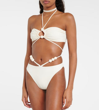 Woman wearing a pearl-accented ivory bikini set with criss-cross halter straps and high-cut bottoms in a tropical garden setting