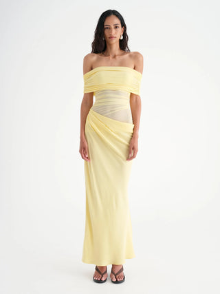 Model wearing buttercream yellow off-shoulder maxi dress with ruched mesh bodice, cowl detail at waist, and flowing overlapping skirt, standing on rocky shoreline with ocean backdrop