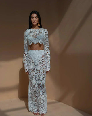 A woman wearing a white crochet floral two-piece beach dress featuring a long-sleeve crop top and a semi-sheer maxi skirt, styled in a bohemian Ibiza beach club aesthetic against a warm-toned background. Vienna Fashion