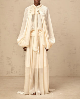 Model wearing ivory satin pleated maxi dress with tie-neck and long sleeves – modest bridal gown or elegant wedding guest dress.
