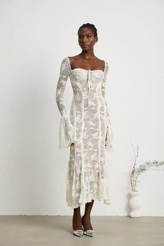 Elegant woman wearing a white bohemian lace maxi dress with long bell sleeves, intricate floral lace pattern, and a sweetheart neckline, standing gracefully in a minimalistic setting with a small vase of flowers. - Cielie Vienna