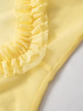 Elegant yellow beach dress with transparent mesh fabric and ruffled details, styled as a bikini cover-up on the grass at a resort.
