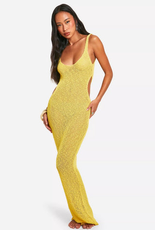 Woman in yellow knit maxi dress with drink, standing by beachside railing under palm trees.