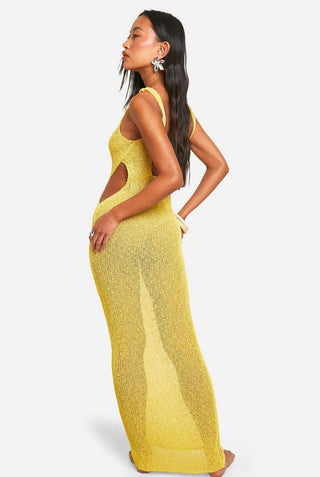 Woman in yellow knit maxi dress with drink, standing by beachside railing under palm trees.