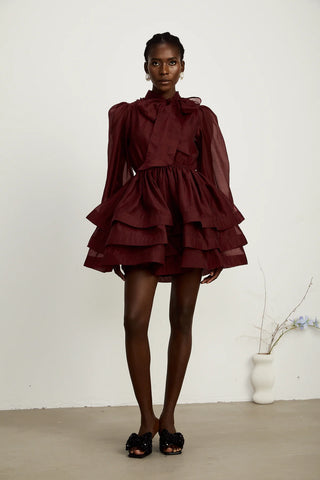 Close-up of a burgundy cocktail dress showcasing pleated details, a dramatic scarf collar with a bow, and semi-sheer fabric for a sophisticated look.