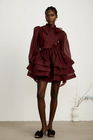 Model wearing a burgundy mini flared dress with tiered layers, long sheer sleeves, and a scarf collar tied into a bow. Perfect for cocktail parties and formal occasions.