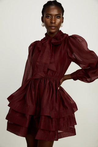 Model wearing a burgundy mini flared dress with tiered layers, long sheer sleeves, and a scarf collar tied into a bow. Perfect for cocktail parties and formal occasions.