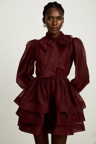 Model wearing a burgundy mini flared dress with tiered layers, long sheer sleeves, and a scarf collar tied into a bow. Perfect for cocktail parties and formal occasions.