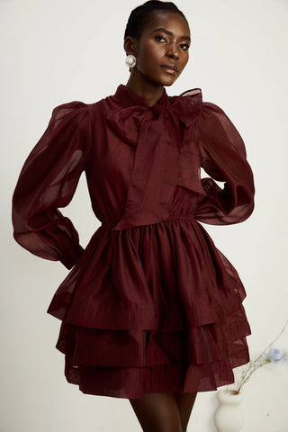 Model wearing a burgundy mini flared dress with tiered layers, long sheer sleeves, and a scarf collar tied into a bow. Perfect for cocktail parties and formal occasions.