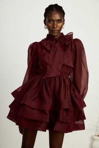 Close-up of a burgundy cocktail dress showcasing pleated details, a dramatic scarf collar with a bow, and semi-sheer fabric for a sophisticated look.