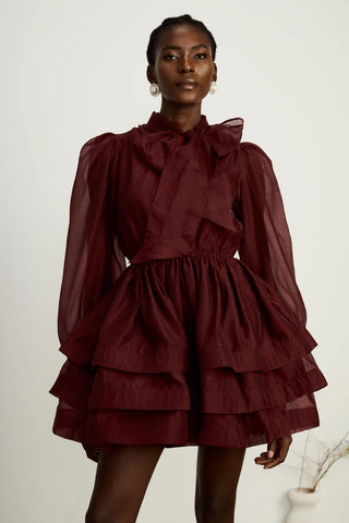 Model wearing a burgundy mini flared dress with tiered layers, long sheer sleeves, and a scarf collar tied into a bow. Perfect for cocktail parties and formal occasions.