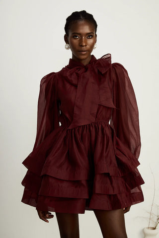 Model wearing a burgundy mini flared dress with tiered layers, long sheer sleeves, and a scarf collar tied into a bow. Perfect for cocktail parties and formal occasions.