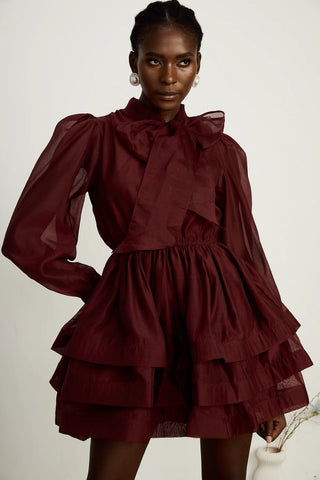 Model wearing a burgundy mini flared dress with tiered layers, long sheer sleeves, and a scarf collar tied into a bow. Perfect for cocktail parties and formal occasions.