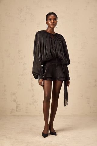 Model wearing the Evelyn Black Draped Mini Dress, showcasing the fully pleated design, twist waist detail, and elegant long buttoned-cuff sleeves.Black Little Dress Ruffled Mini Longsleeve