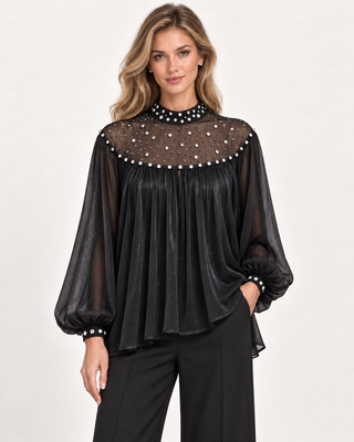 A woman modeling a black sheer long-sleeve blouse with a high mock neck, embellished with pearl-like bead accents around the neckline and cuffs, paired with black trousers against a plain light background.