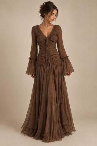A woman modeling a floor-length brown gown with long flared sleeves, a deep V-neckline, gathered ruching at the bodice, and a flowing pleated skirt against a plain beige studio background.