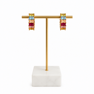 A pair of gold drop earrings with stacked rectangular gemstones in light blue, pink, and deep red, displayed on a gold T-stand with a white marble base against a white background.