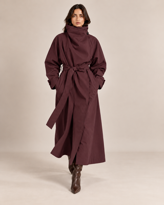 A model wearing a long, dark burgundy wrap coat with a high folded collar and waist tie, paired with brown leather boots, against a solid beige studio background.