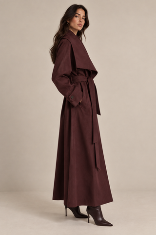 A woman modeling a long, dark brown wrap-style coat with a belted waist, shown in profile against a plain beige studio background.