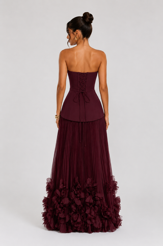 A woman modeling a strapless floor-length gown from the back, featuring a structured lace-up corset bodice, a pleated skirt, and a ruffled tulle hem in deep burgundy, against a light gray studio background.