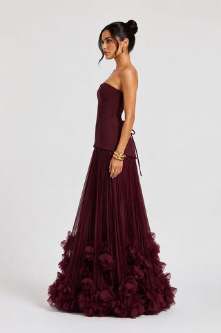 A woman modeling a strapless floor-length gown in deep burgundy. The dress features a structured bodice, a flowing pleated skirt, and a tiered ruffle hem with matching fabric flowers.