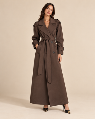 A woman modeling a long brown trench coat with a matching waist tie, photographed against a plain beige studio background.