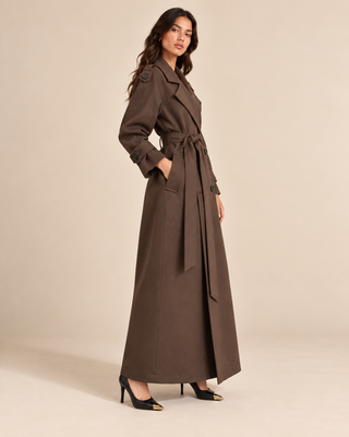 A woman modeling a long brown trench coat with a matching waist tie, photographed against a plain beige studio background.