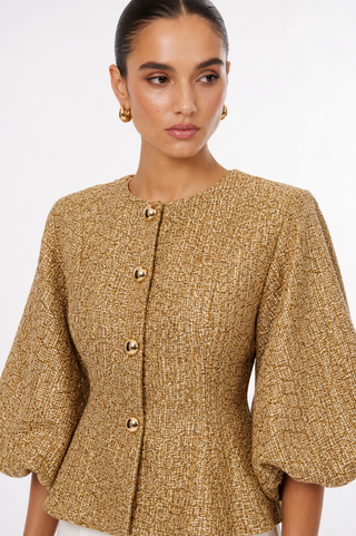 A woman wearing a textured gold tweed jacket with puff sleeves and gold buttons, photographed against a clean white studio background.