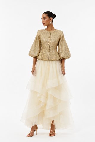 A model wearing a gold sequined jacket with puff sleeves and a long cream tulle ruffled skirt, standing against a plain white studio background.