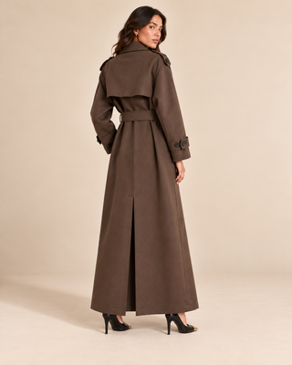 A woman modeling a long brown trench coat from the back, standing against a plain beige studio backdrop.