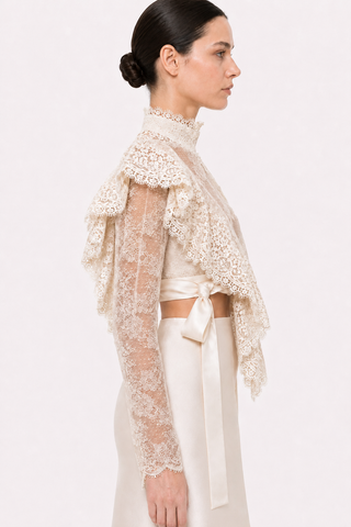 A woman shown in side profile wearing a cream lace long-sleeve shrug or bolero over a matching cream satin top and skirt, against a plain light background.