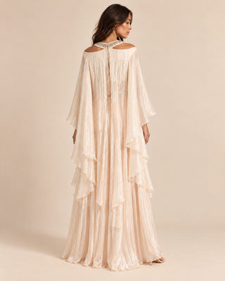 A woman modeling a floor-length cream or off-white maxi dress with a backless open-shoulder design, layered ruffle hem, and sheer long sleeves, shown from the back against a warm beige studio background.