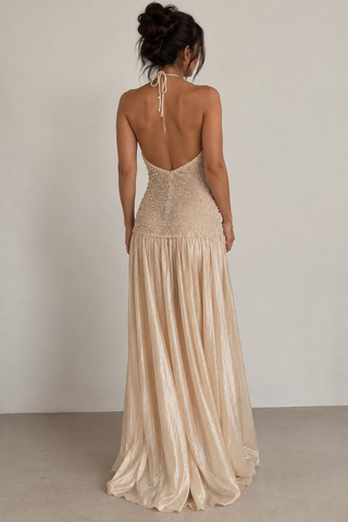A woman viewed from the back wearing a champagne-colored, sleeveless halter-neck maxi dress with a beaded open back and a flowing pleated skirt, set against a neutral studio background.