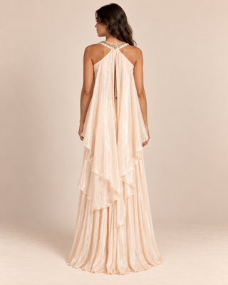 A woman modeling a floor-length halter gown from the back, shown against a plain warm beige studio background. The dress has a sheer draped overlay with a lace-up open back and a layered ruffle effect.
