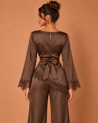 A woman shown from the back wearing a brown satin jumpsuit with lace-trimmed cuffs and a matching waist tie, against a solid warm tan studio background.