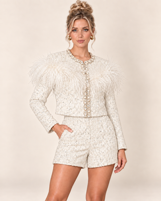 A woman models a matching cream tweed jacket and shorts set with feathered shoulder details and pearl-like embellishments, against a solid beige studio background.