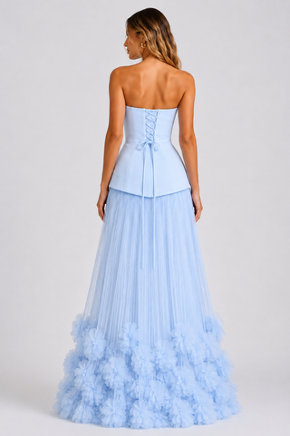 A woman shown from the back wearing a strapless light blue formal gown with a structured lace-up corset-style bodice and a voluminous tulle skirt with layered ruffle details near the hem, photographed against a plain light gray studio background.