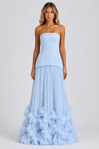 A woman modeling a strapless light blue gown with a fitted bodice and a voluminous pleated skirt that ends in layered ruffles, photographed against a plain light gray studio background.