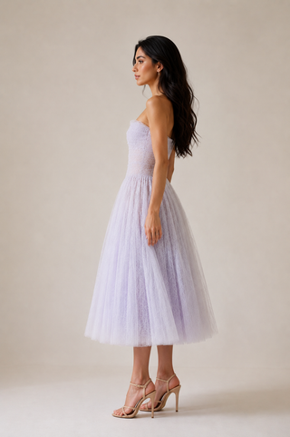 A woman modeling a strapless lavender tulle midi dress with a fitted bodice and full skirt, paired with nude high-heeled sandals, against a plain beige studio background.