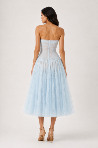 A woman shown from the back wearing a strapless light blue tulle dress with a structured corset-style bodice and a full midi skirt, paired with nude high heels, against a plain light gray studio background.
