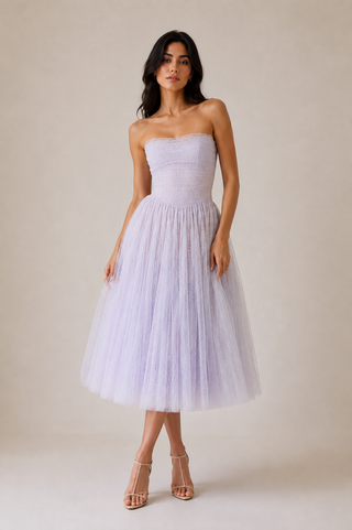 A woman modeling a strapless lavender tulle midi dress with a fitted bodice and full skirt, paired with strappy high-heeled sandals, against a plain beige studio background.