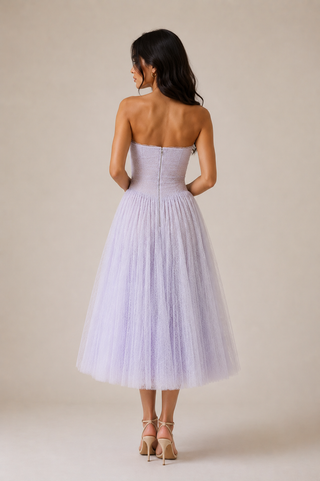 A woman stands with her back to the camera wearing a strapless lavender tulle midi dress with a fitted corset-style bodice and a full layered skirt, paired with beige high-heeled sandals against a plain beige studio background.