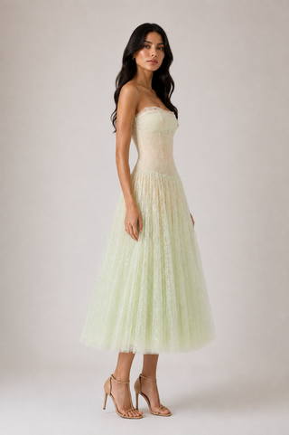 A woman modeling a strapless pale yellow-green tulle midi dress with a fitted lace bodice, paired with nude high-heeled sandals, against a plain light beige studio background.