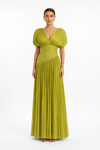 A woman modeling a floor-length olive green gown with voluminous short puff sleeves, a deep V-neckline, a fitted waist, and a pleated skirt, photographed against a plain white studio background.