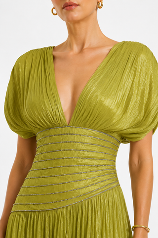 A close-up of a model wearing a shimmering olive green dress with a deep V-neckline, puffed short sleeves, and a heavily embellished ruched waistband with horizontal rows of sparkling crystals.