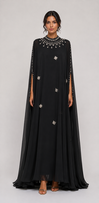 A woman modeling a long black floor-length gown with a draped cape-like overlay, embellished with silver beaded details around the high neckline and scattered star-shaped motifs on the skirt, against a light gray studio background.